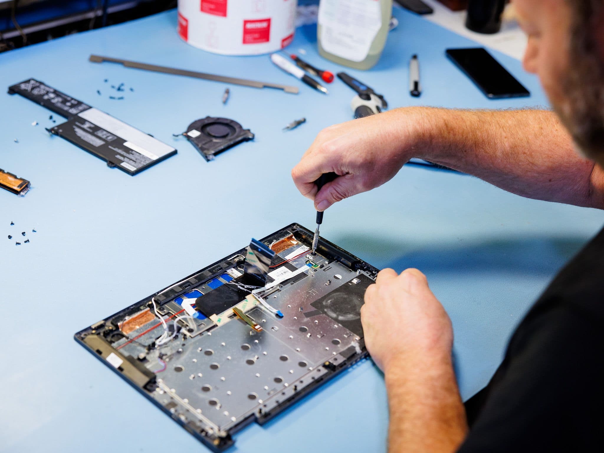 A person's hands repairing an open laptop with a screwdriver, surrounded by disassembled components on a blue desk.