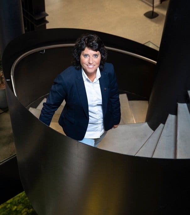 A smiling woman in a navy blazer stands on a modern spiral staircase, looking up.