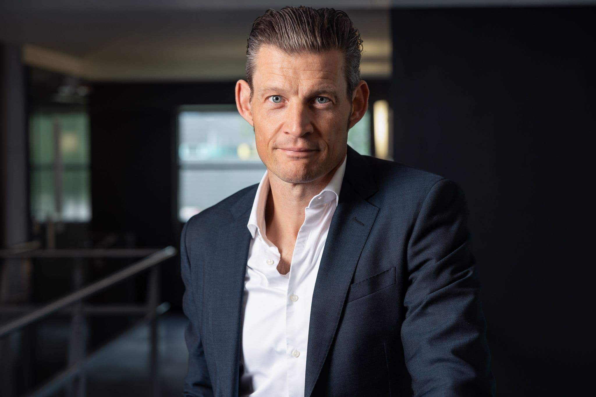 A man in a dark suit jacket and white shirt looks directly at the camera.