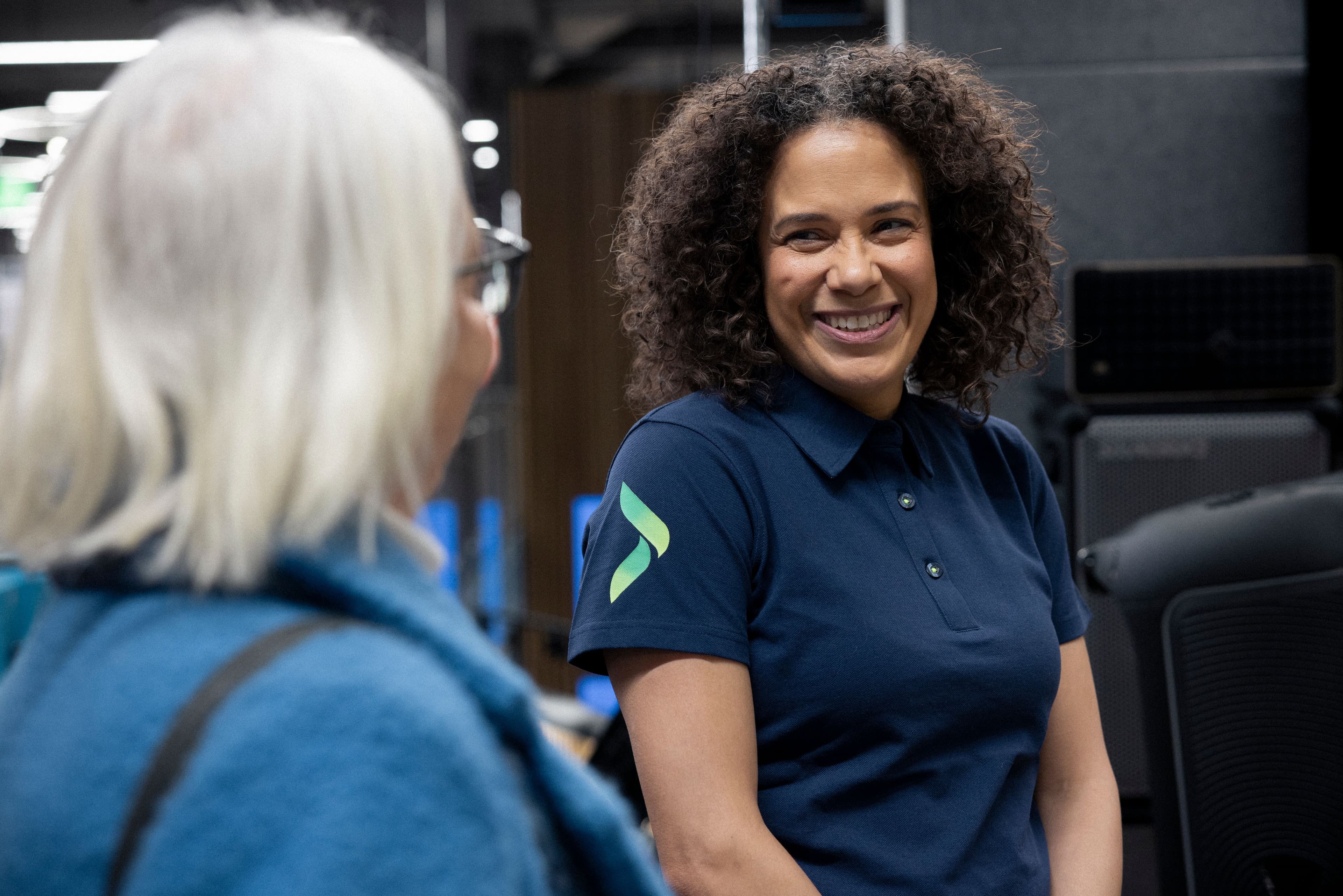 Smiling woman in blue polo with green logo talking to a person with their back turned.