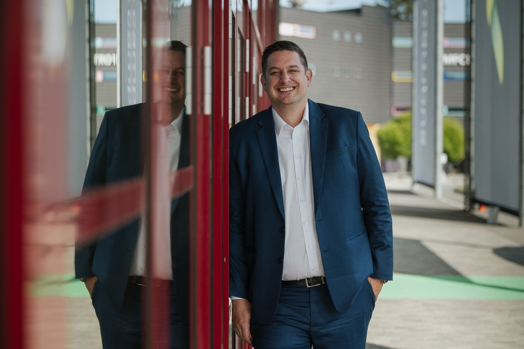 Smiling man in a blue suit leaning on a red wall, with his reflection visible.