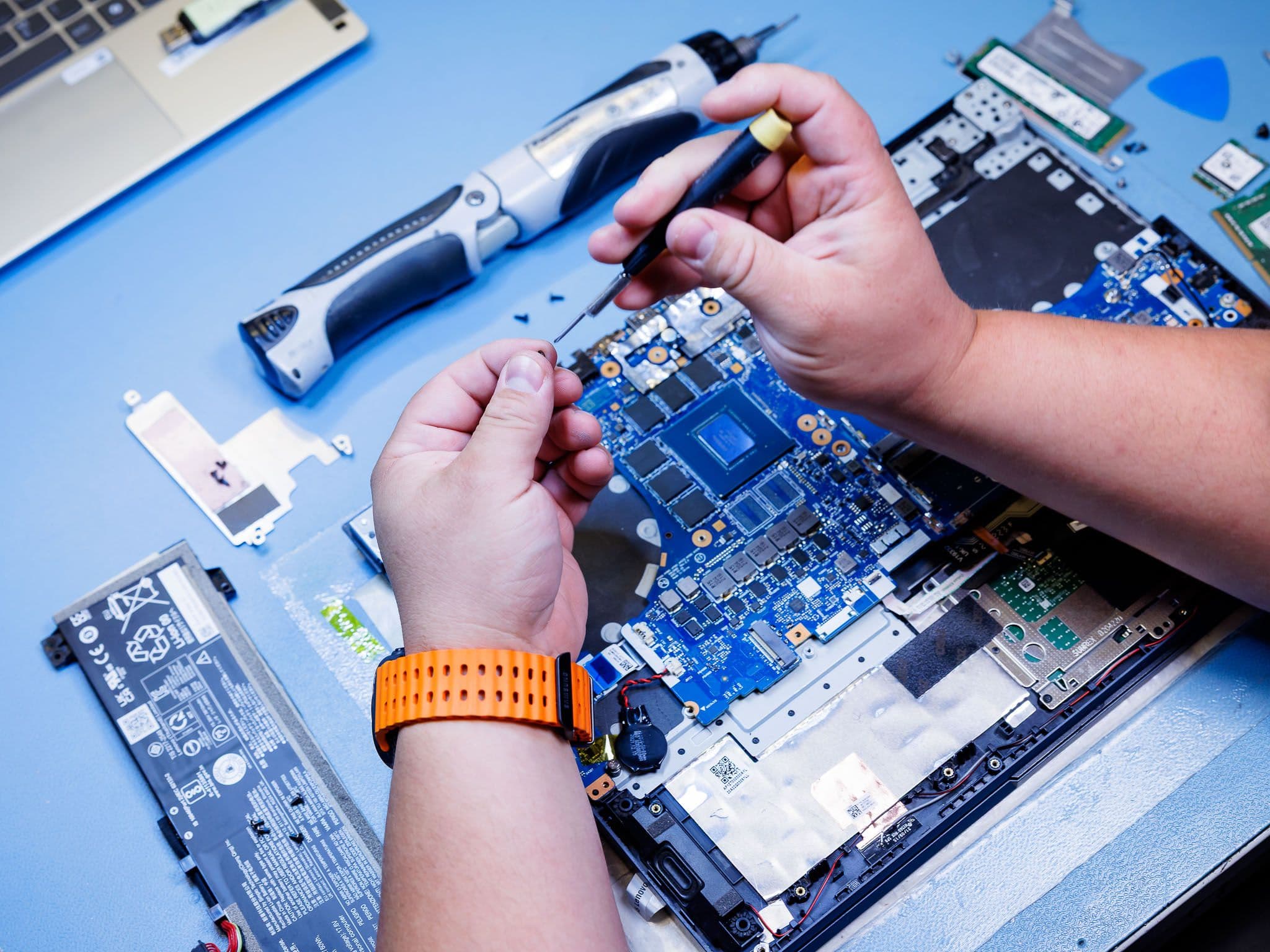 Hands use a small screwdriver to work on a laptop's open motherboard and components on a blue surface.