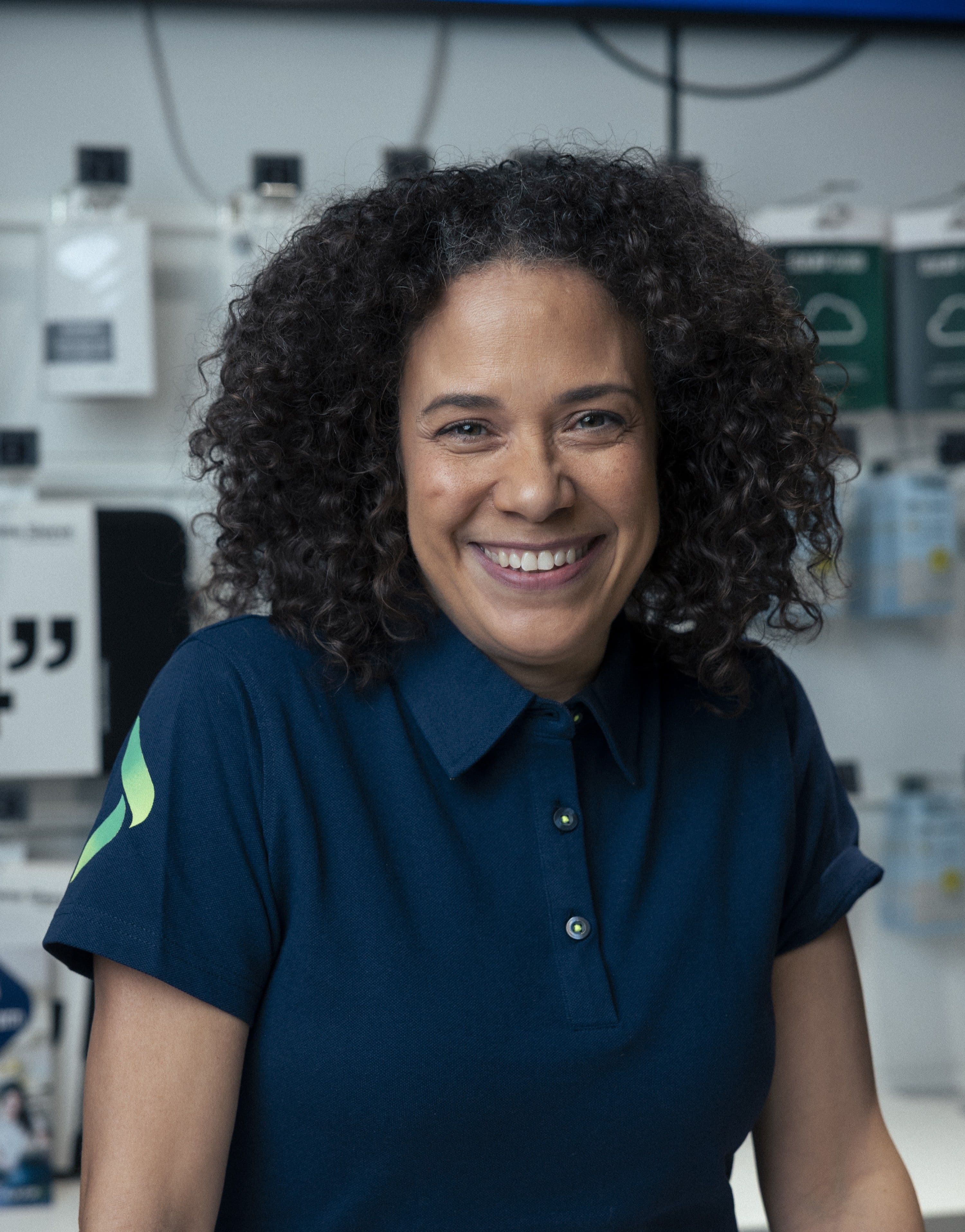 A smiling woman with curly hair wears a dark blue polo shirt with a green and yellow logo on the sleeve.