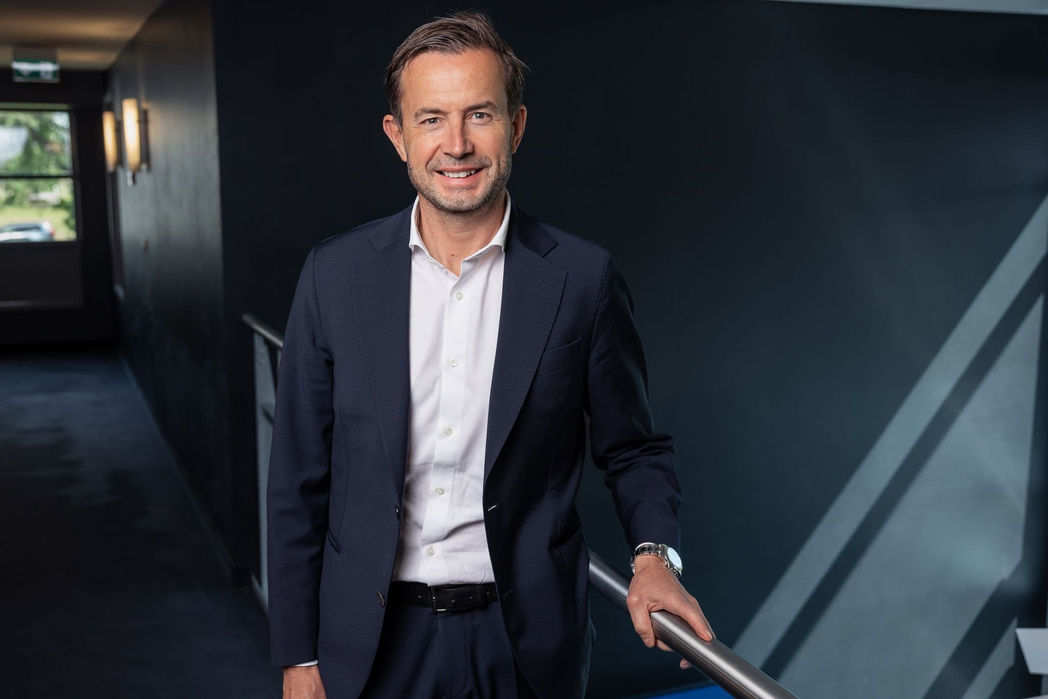 A smiling man in a dark suit holds a handrail on a staircase.